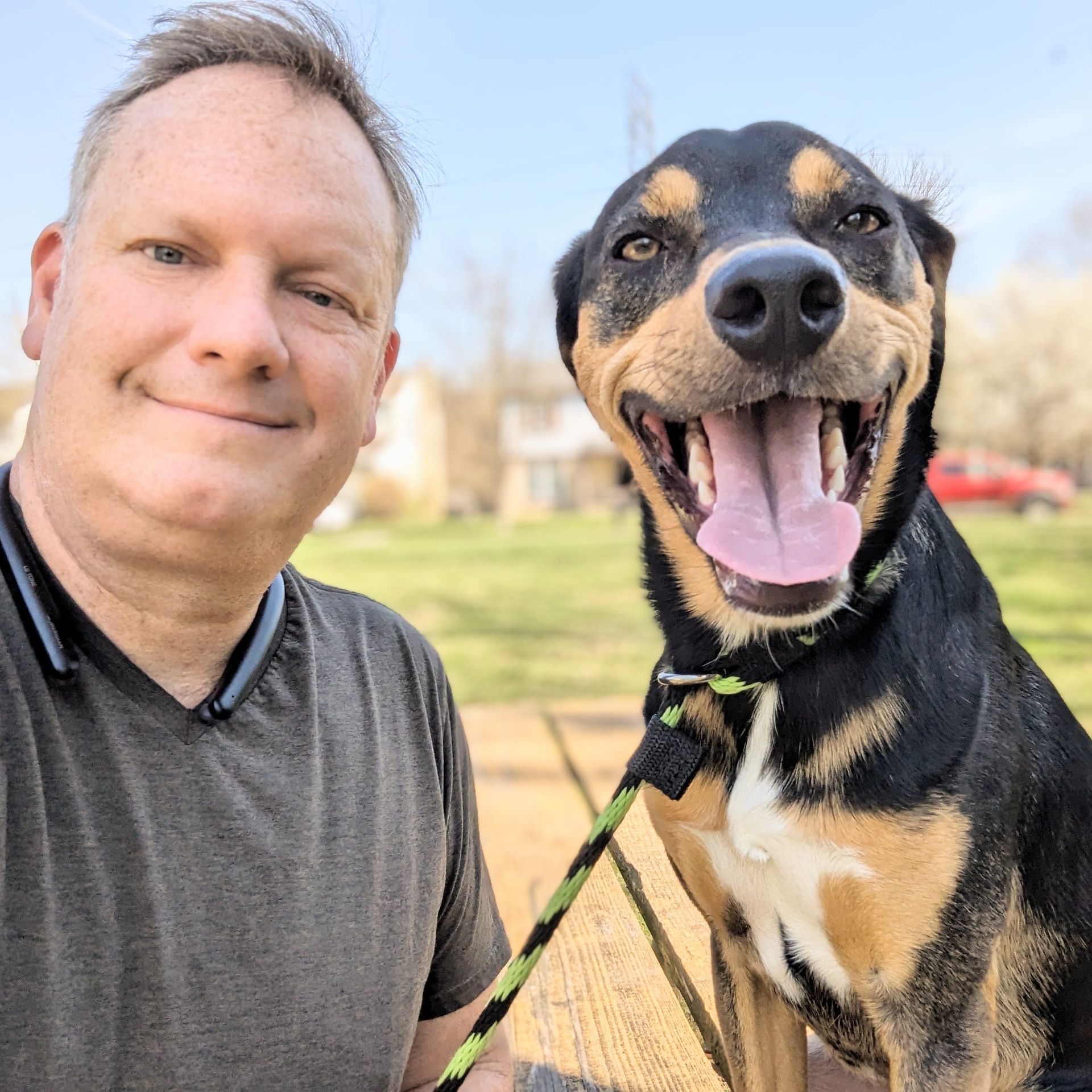 A man poses for a selfie outdoors next to a happy, black and tan dog with its mouth open, wearing a green leash. Dog trainer in Northville MI.