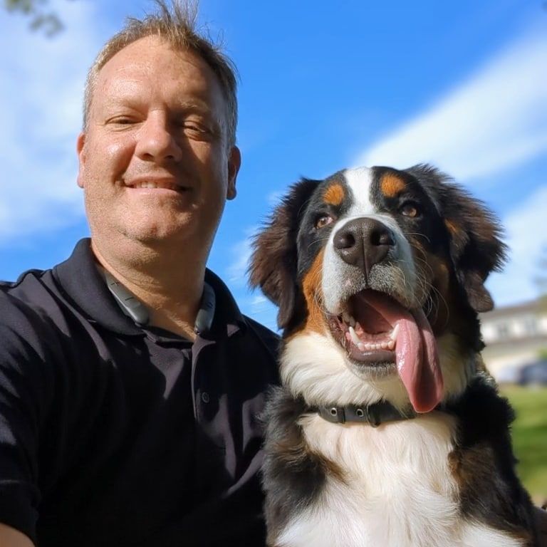 A man smiles while taking a selfie outdoors next to a happy, panting Bernese Mountain dog with its tongue hanging out. Dog trainer in Northville MI. 