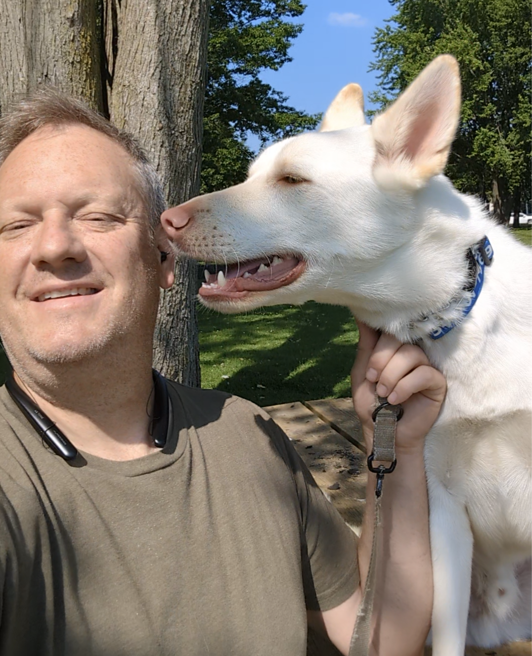 A man wearing a green shirt poses for a selfie outdoors while a white dog nuzzles his cheek. Dog trainer in Ann Arbor MI.