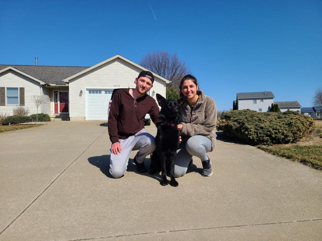Two people kneeling on a driveway with a black dog.