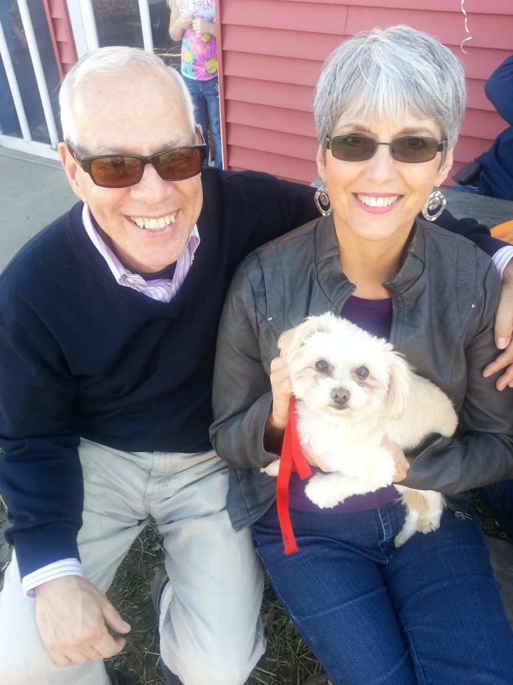 A smiling couple holding a small white dog with a red leash, standing in front of a red building.