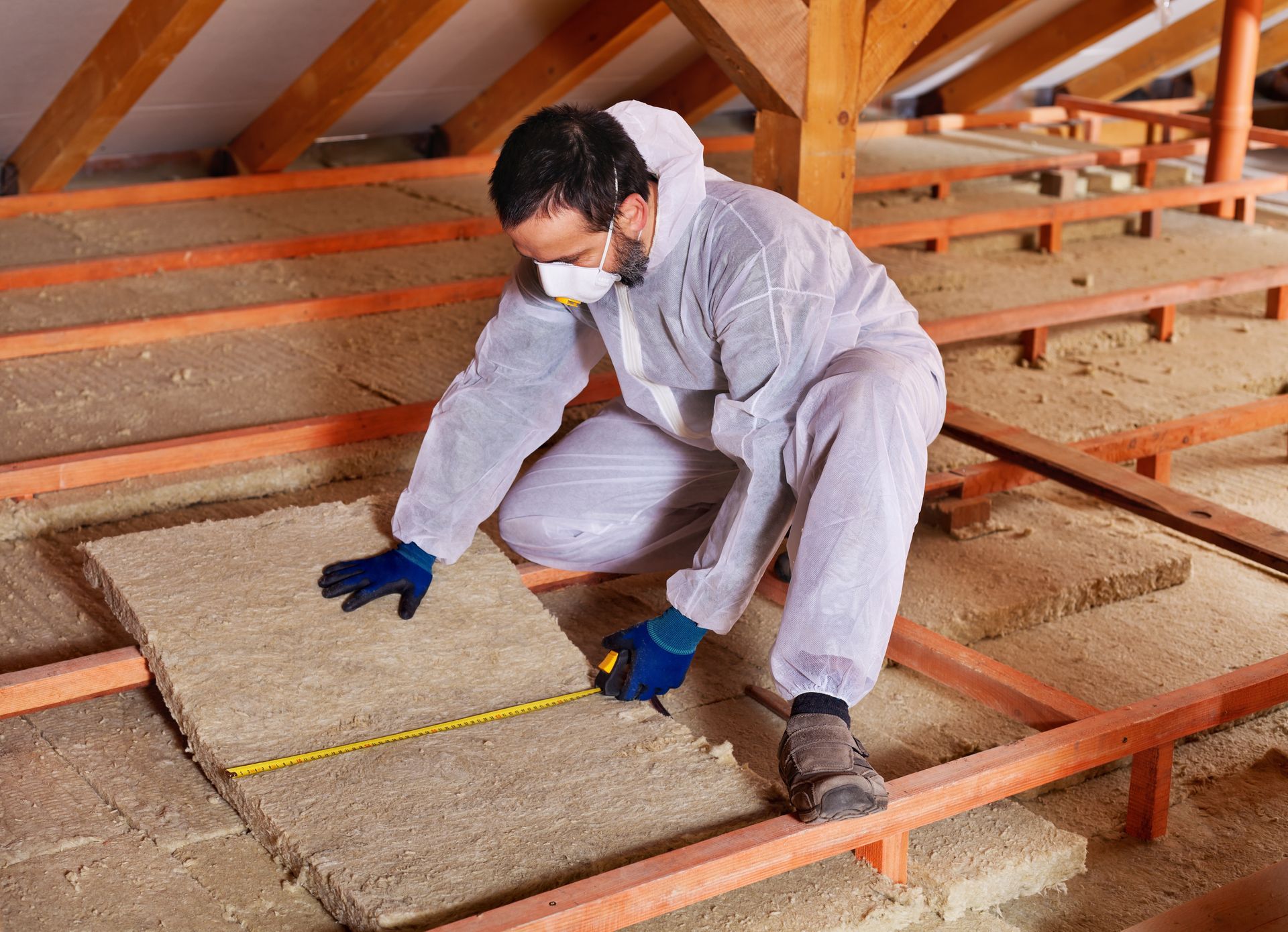 Man in protective suit measuring insulation in an attic, wooden beams visible. Man in protective suit measuring insulation in an attic, wooden beams visible.