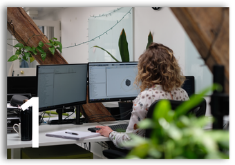 Person working at a desk with dual monitors in a bright office, viewed from behind.
