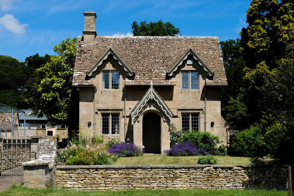 Stone cottage with a steep roof, arched doorway, and purple flowers in front, under a blue sky