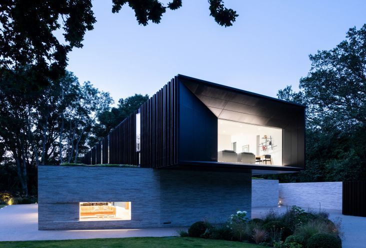 Modern house at dusk with dark siding, lit windows, and trees surrounding it