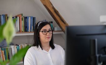 Woman in glasses working at a desk in an office, with bookshelves and a computer monitor nearby.