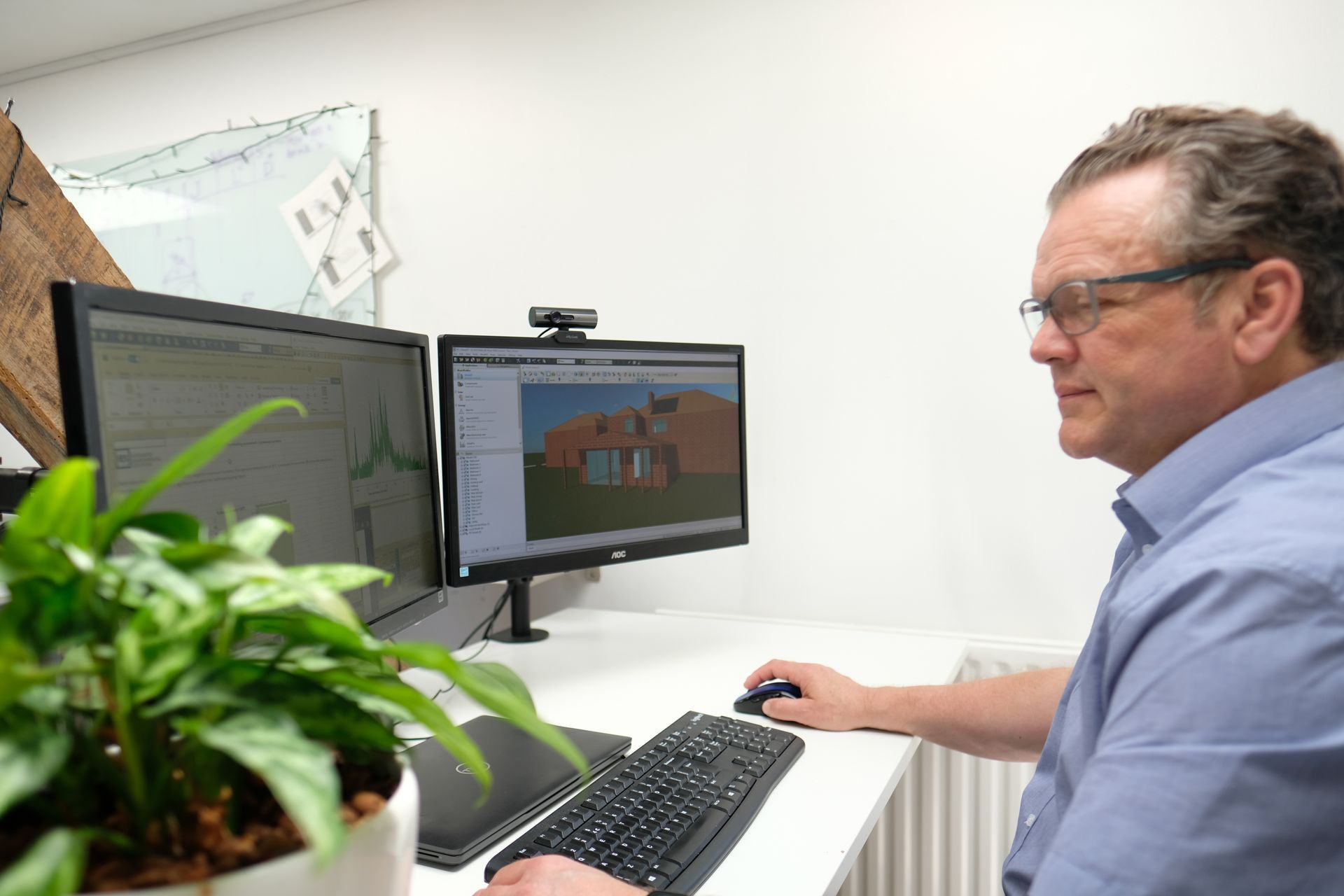 Person working at a desk with dual monitors, editing a 3D house design on screen.