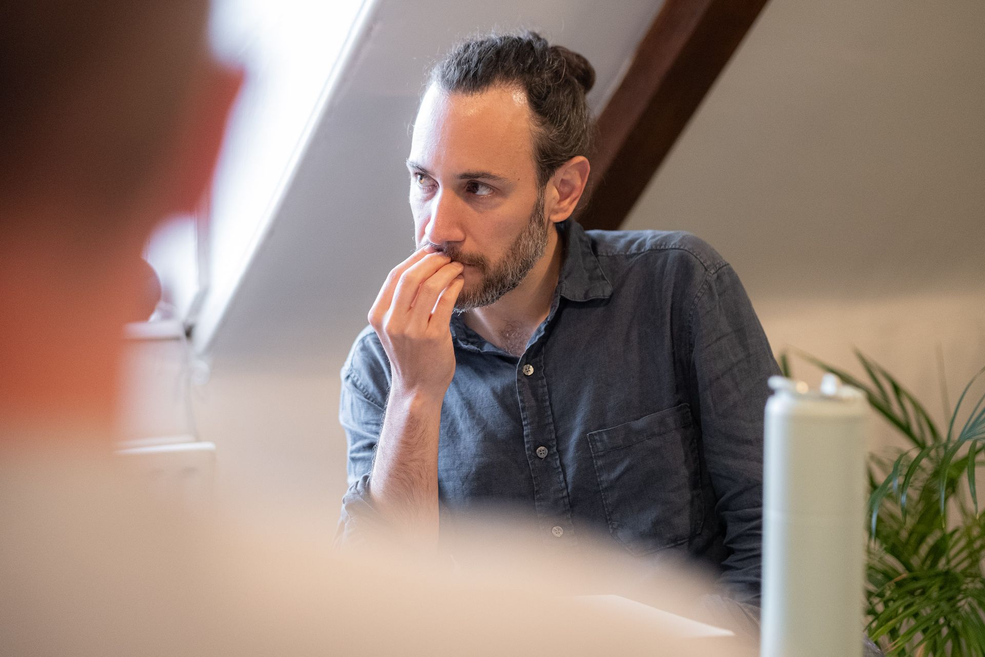 Man sitting at a desk, thinking with hand near mouth, in a bright office.