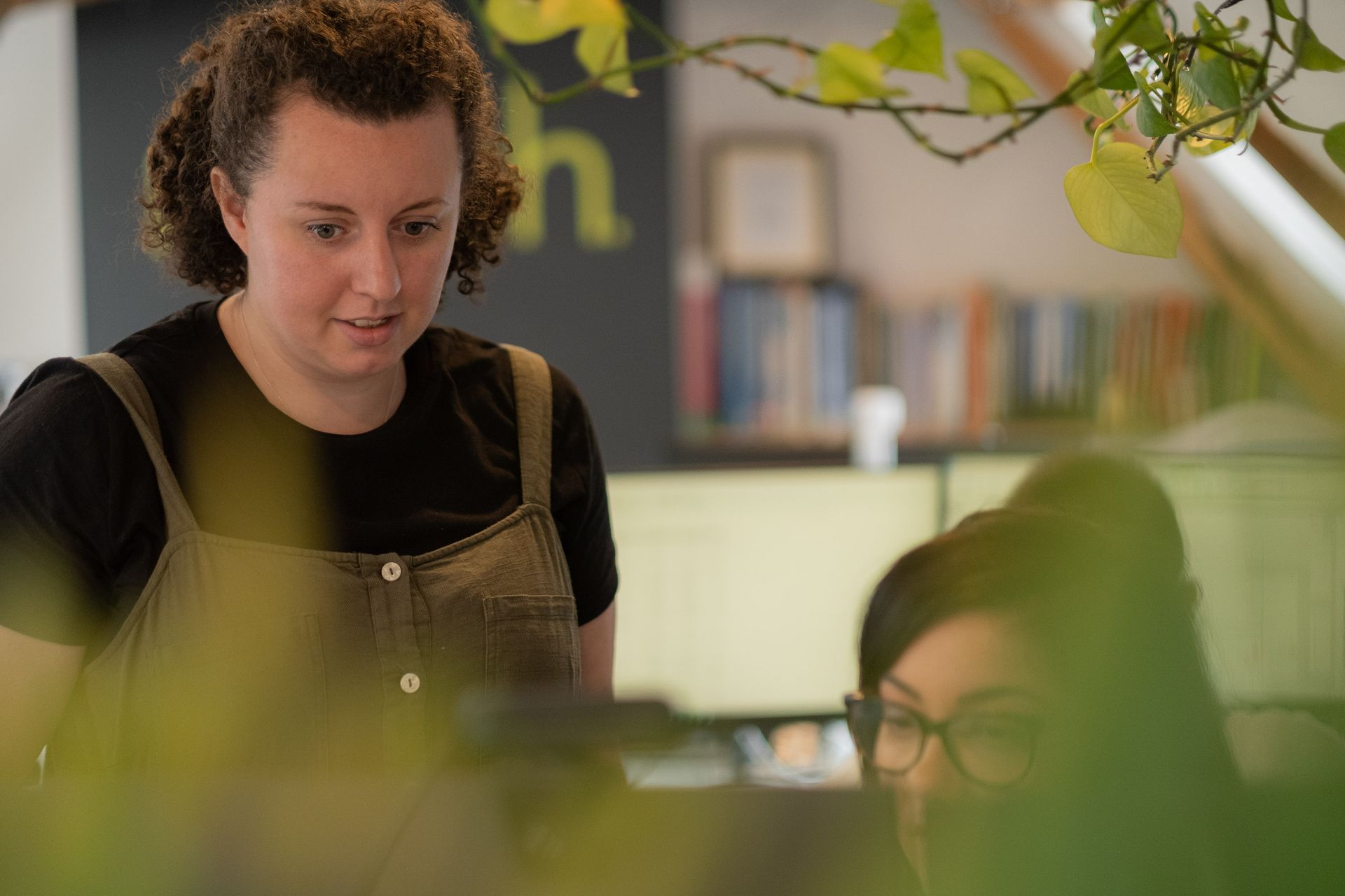 Two people talking in a cozy indoor café, with one smiling behind green plants.