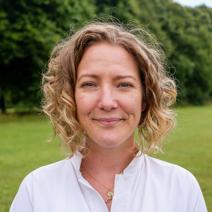 Woman smiling outdoors in a white blouse, with a green lawn and trees in the background