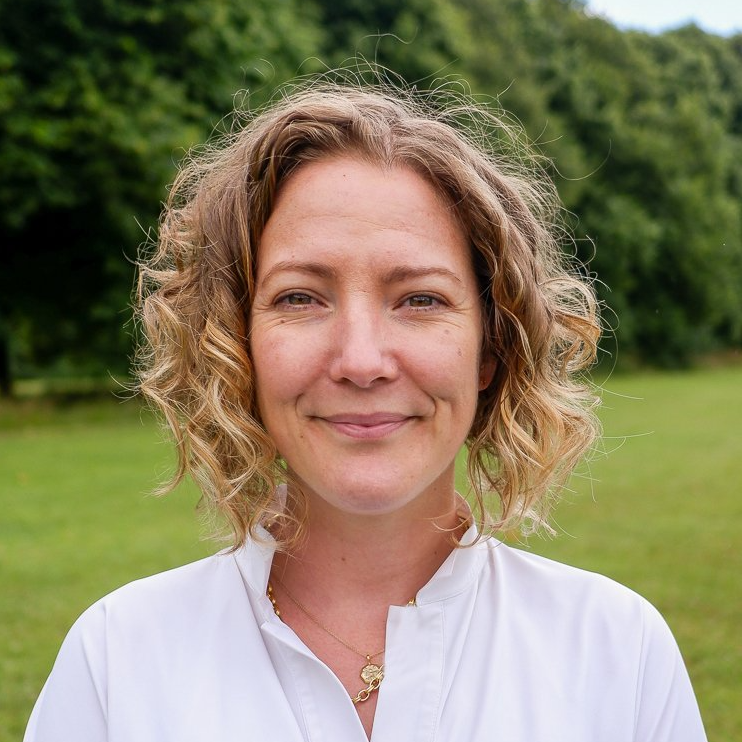 Woman smiling outdoors in a grassy park, wearing a white blouse with curly hair.