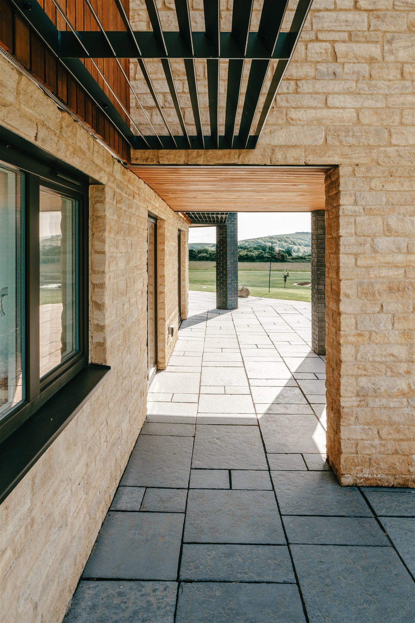 Stone corridor with wooden beams, window on the left, and sunlight leading to a grassy view outdoors