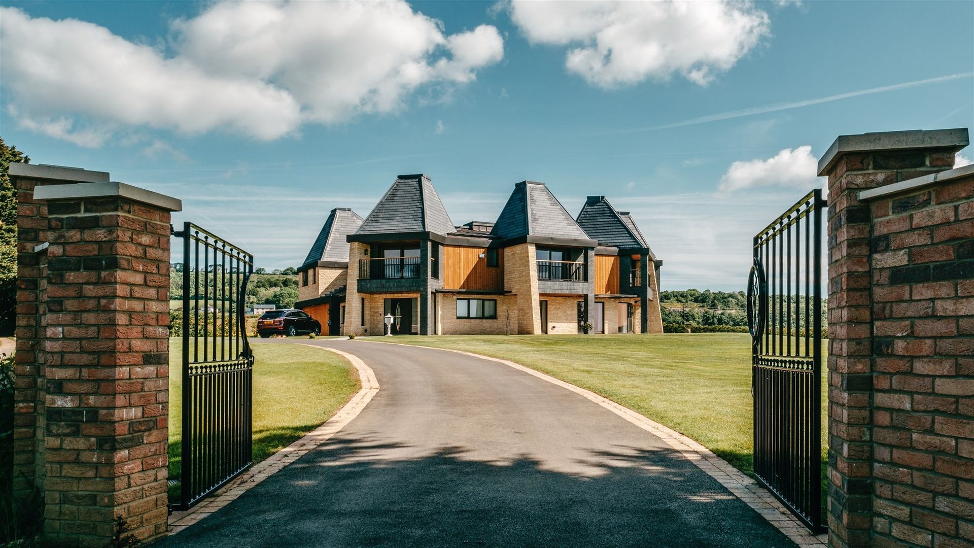 Open wrought-iron gates leading to a large brick house with a curved driveway and lawn under a blue sky