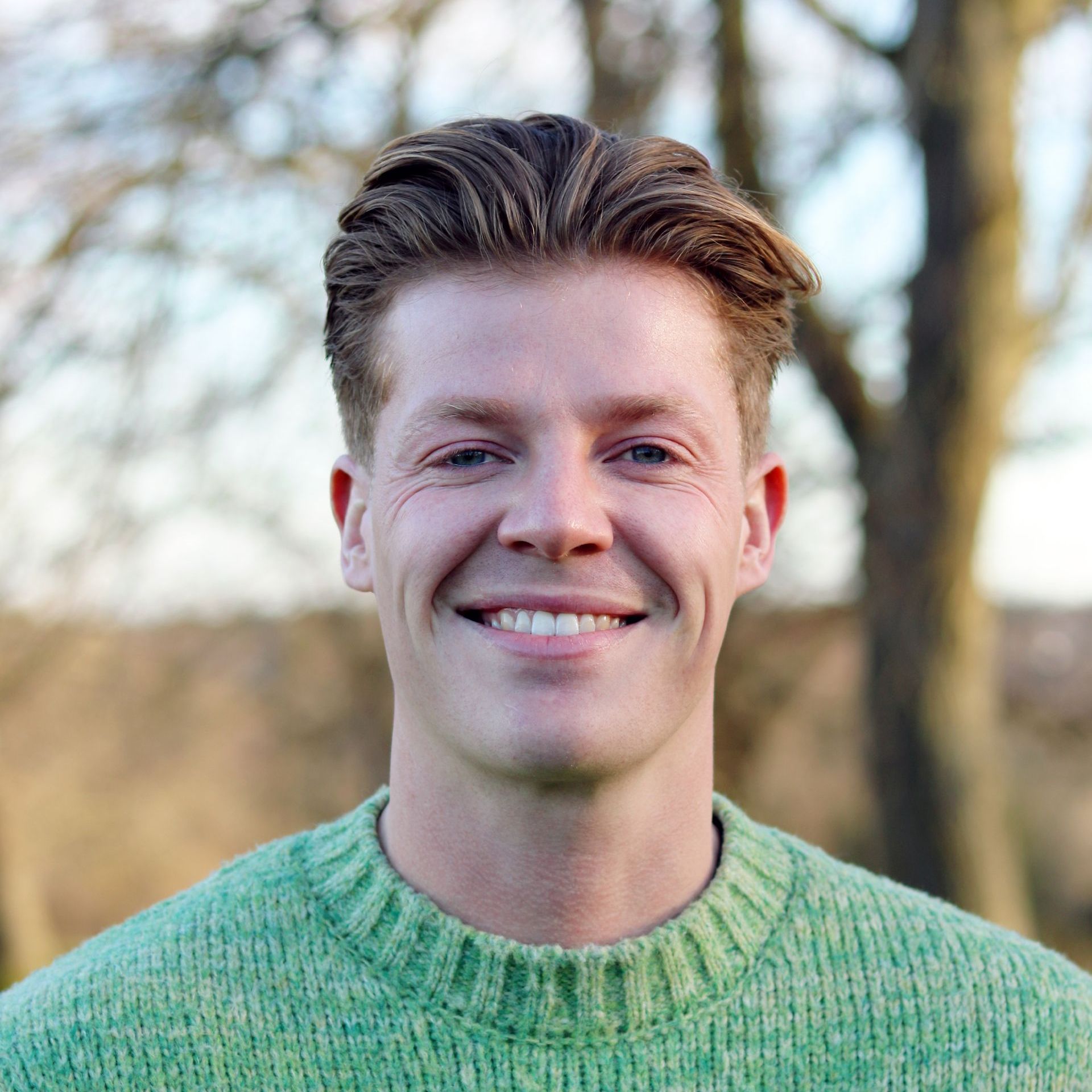 Smiling person in a green sweater outdoors with blurred trees in the background