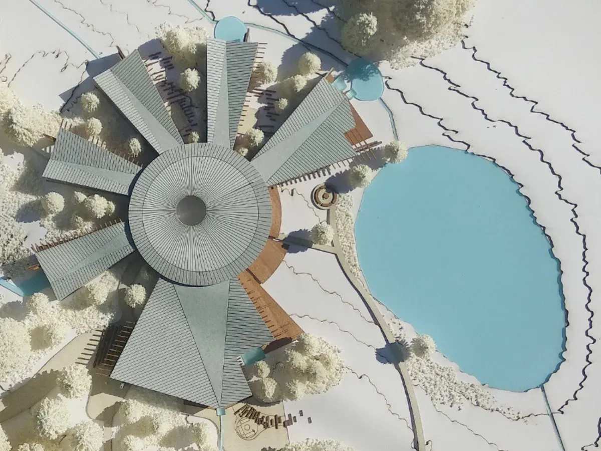Aerial view of a circular building model beside a blue pond, with radiating paths and trees