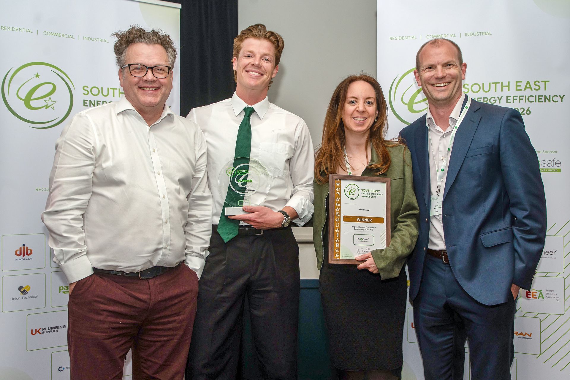 Four people posing at an awards event, one holding a framed certificate, in front of a logo backdrop