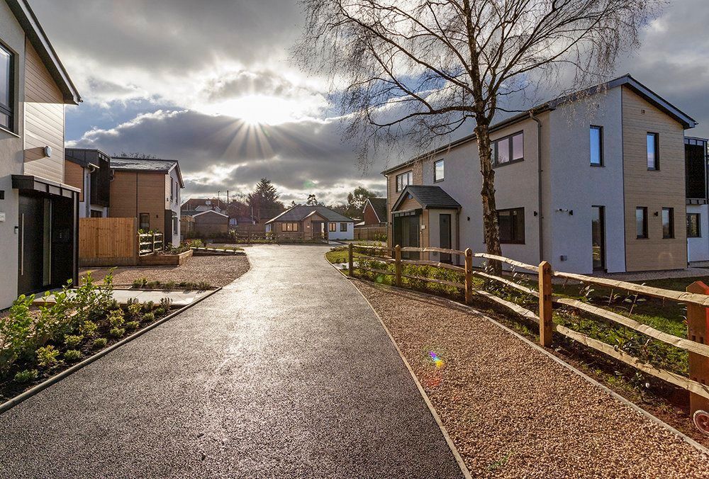 Sunlit residential walkway between modern houses, with trees, fences, and a cloudy sky