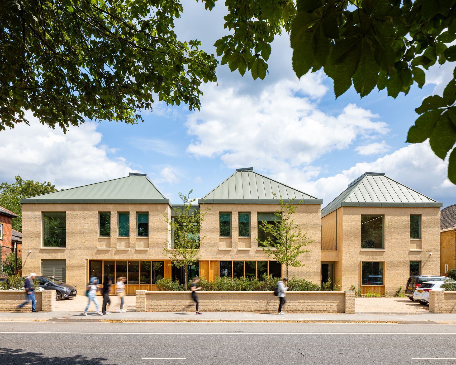 Three tan brick townhouses with green roofs and trees lining the sidewalk on a sunny street.