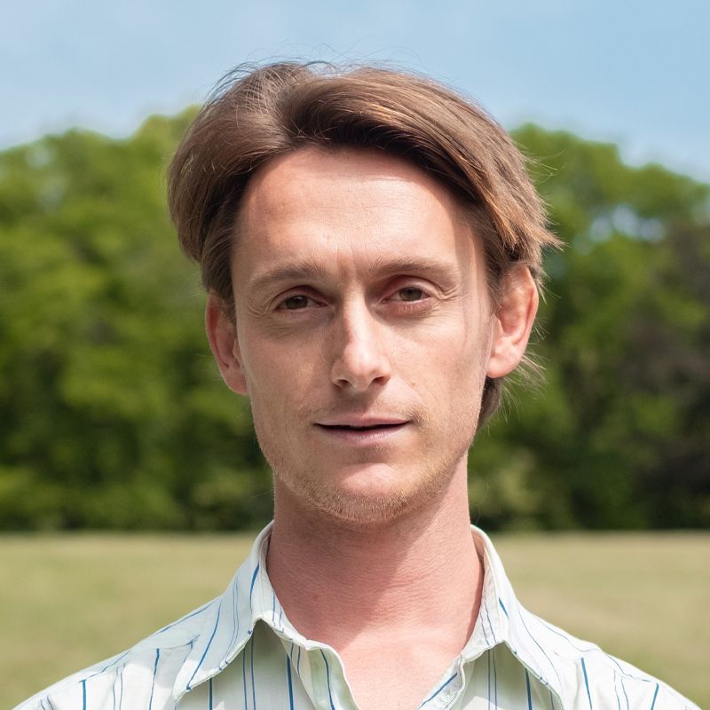 Portrait of a man outdoors in a park, wearing a light striped shirt with trees and grass in the background