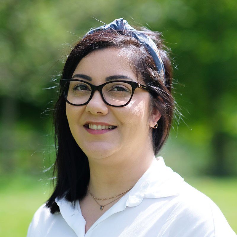 Portrait of a smiling person with black glasses and white shirt outdoors, with green blurred background