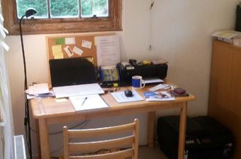 Small desk workspace by a window with papers, laptop, mug, and a corkboard above it.