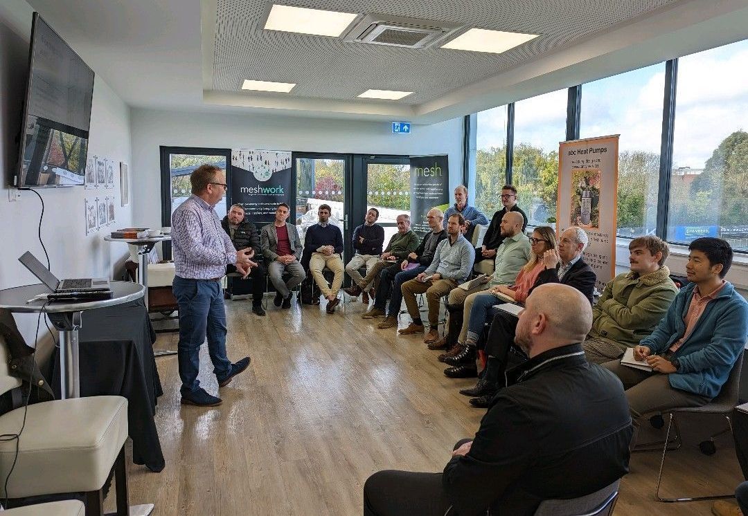 Presenter speaking to a seated audience in a bright room with large windows.