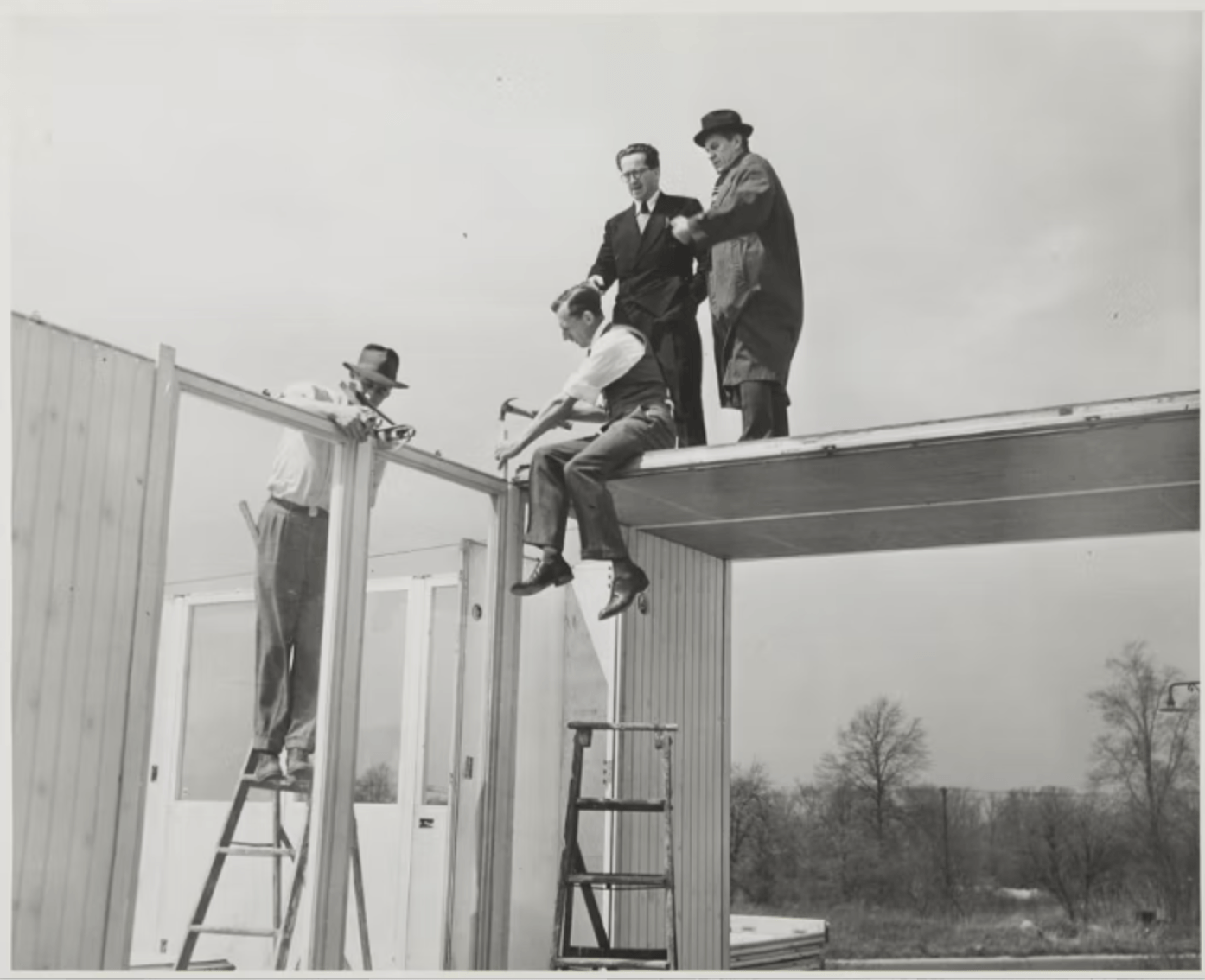 Men on ladders and a roof frame, building a house exterior in black and white.
