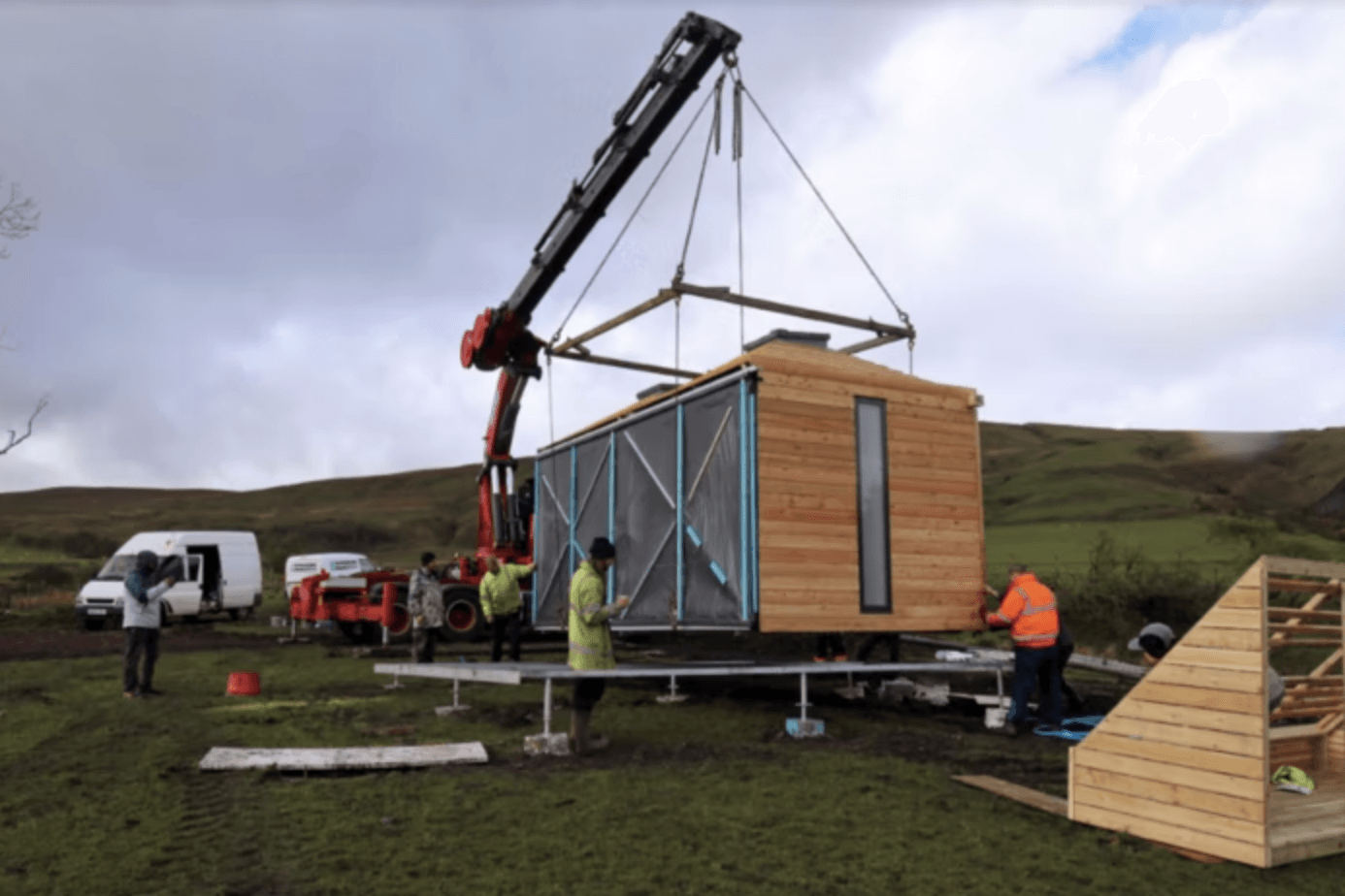 Crane lifting a small wooden cabin onto a platform in a grassy field with workers nearby