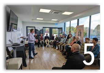 Meeting room with people seated in a semicircle while a speaker stands at the front near a presentation screen