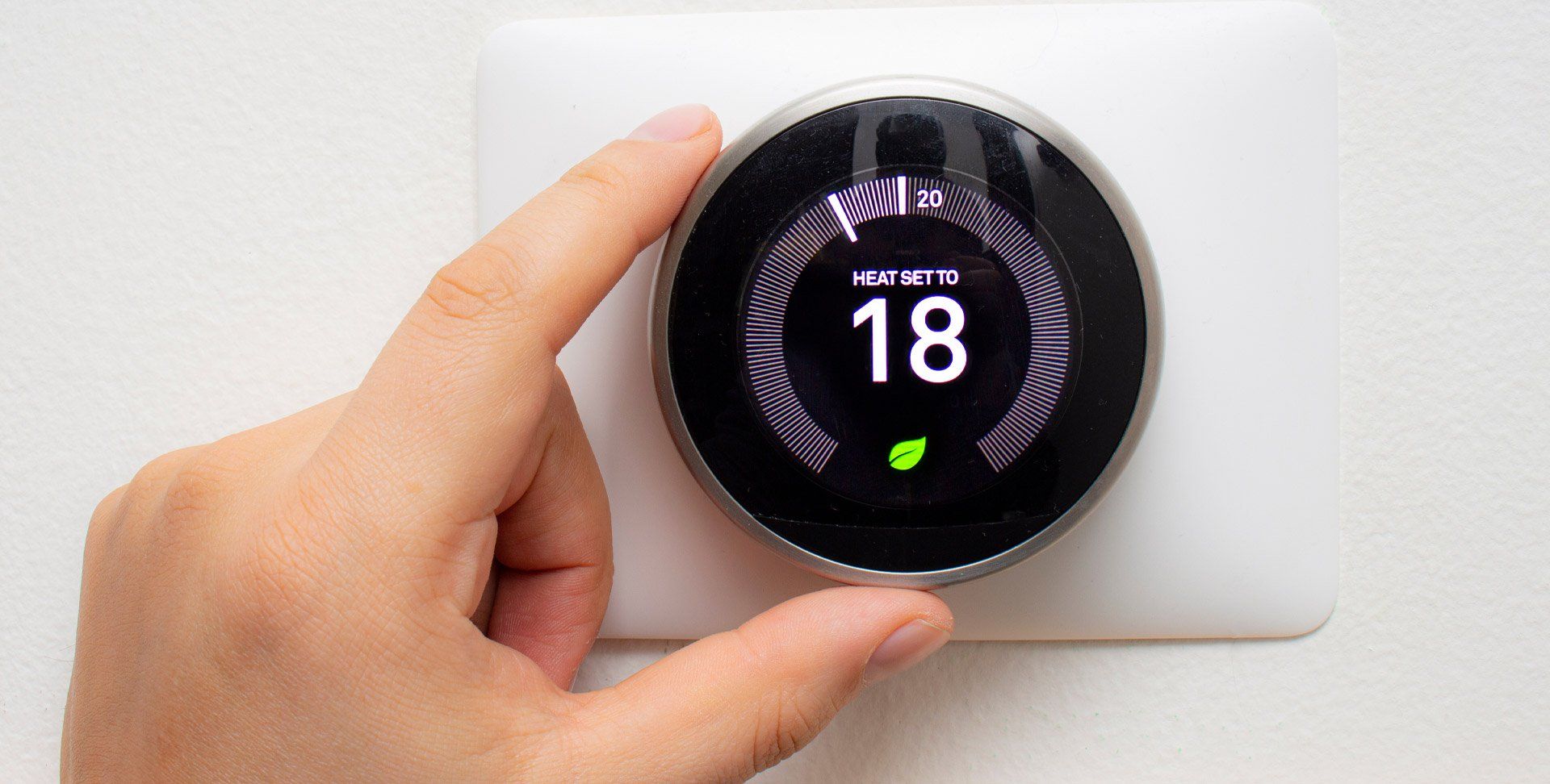 Hand adjusting a round smart thermostat on a white wall, showing 18°