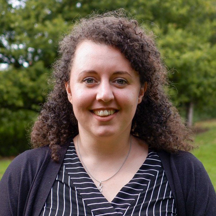 Smiling person with curly hair wearing a striped shirt and dark cardigan outdoors with trees behind them