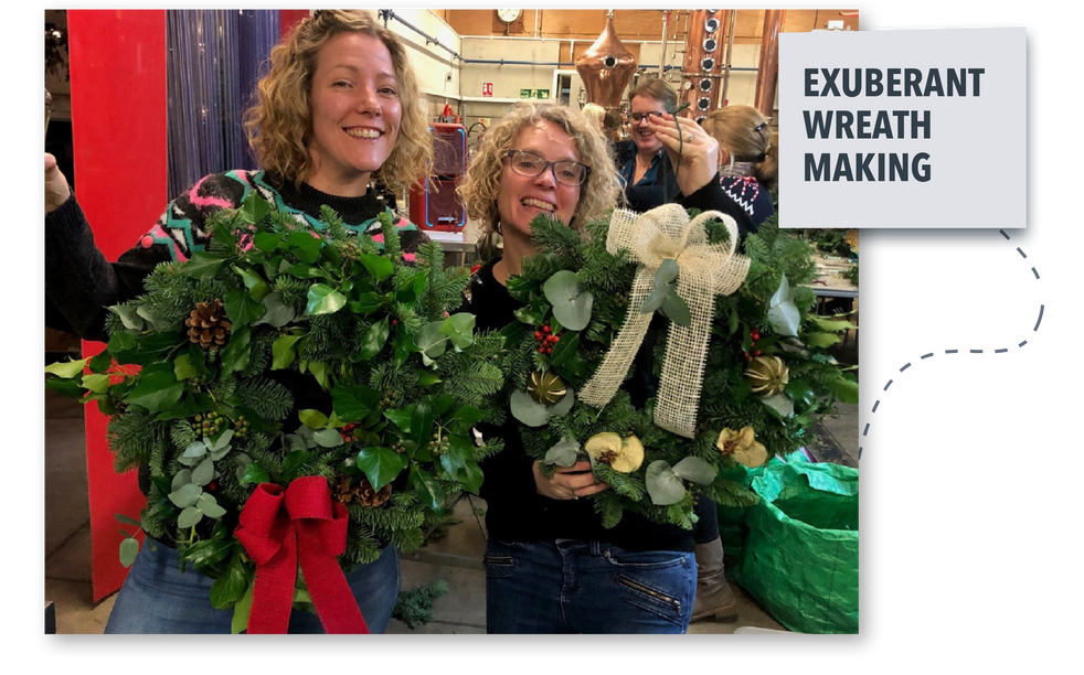 Two women smiling while making large decorative wreaths in a shop, with a sign reading “Exuberant Wreath Making”