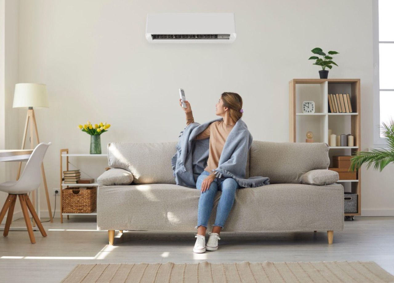 Woman sitting on a beige sofa in a bright living room, pointing at an air conditioner on the wall.
