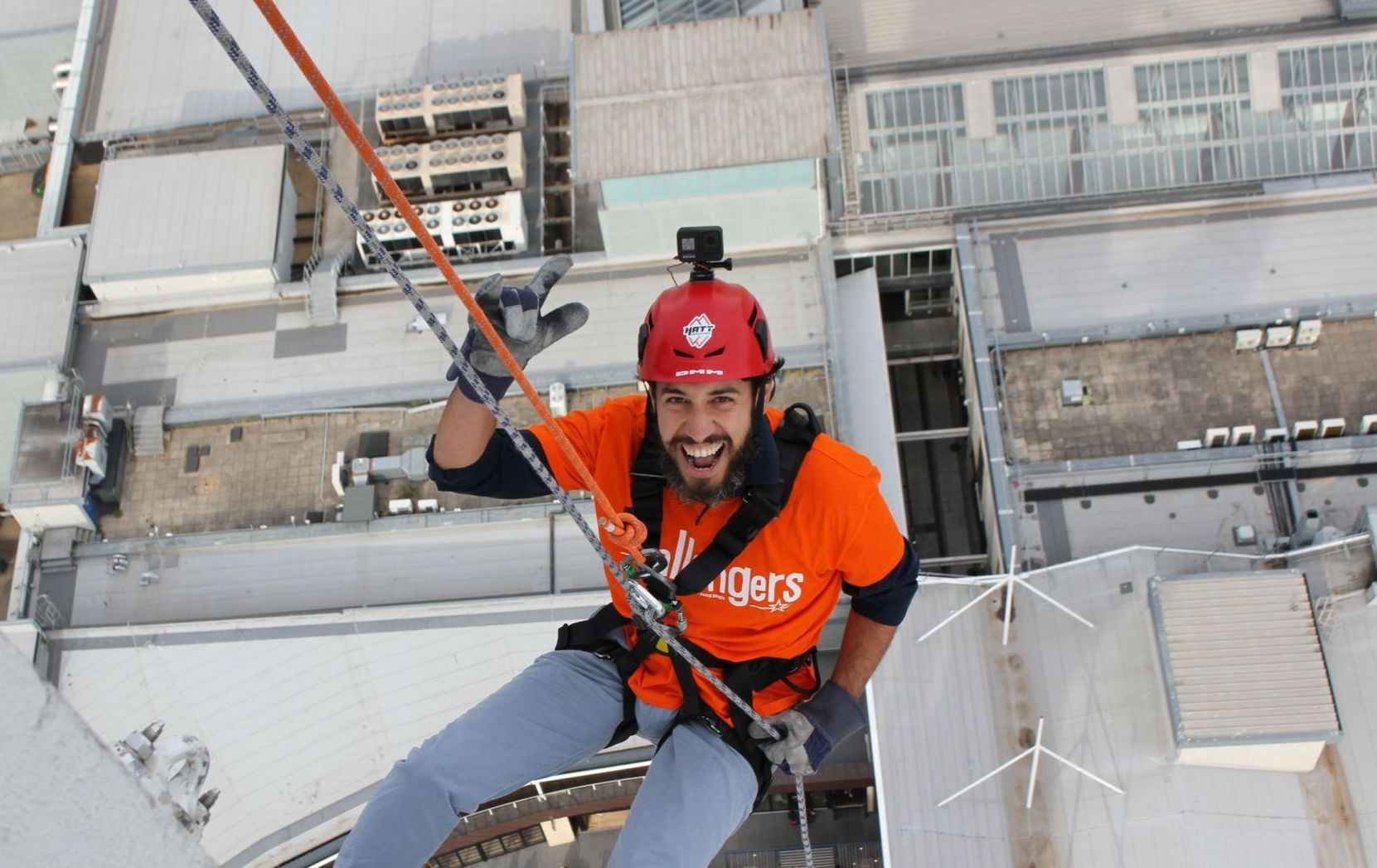 Worker in orange safety gear rappelling from a rooftop above city buildings