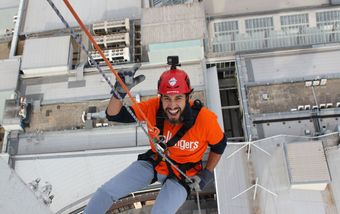 Worker in orange safety gear rappelling from a rooftop above city buildings