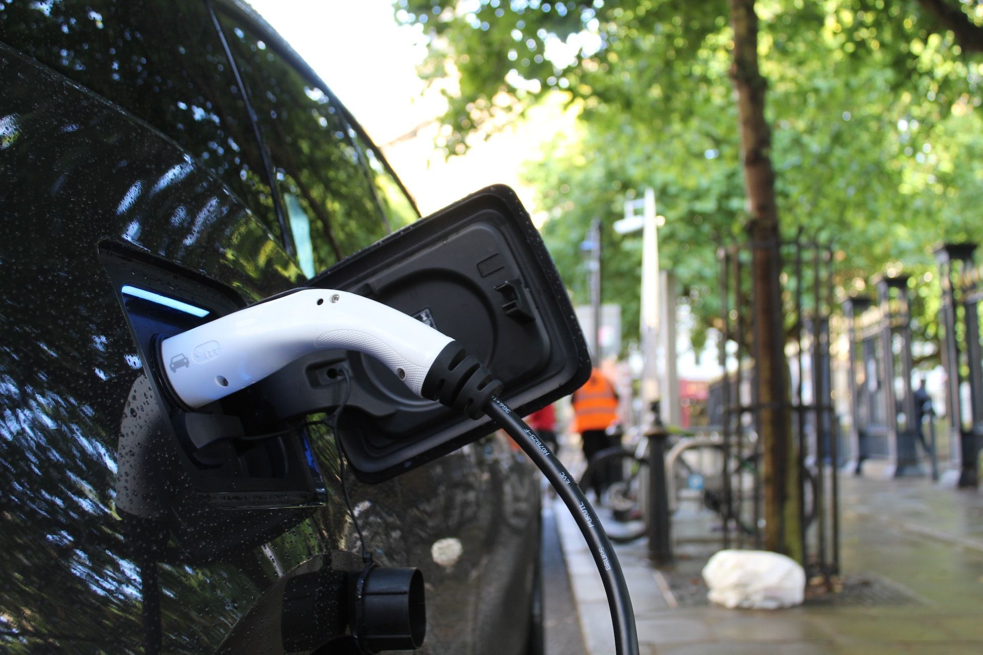 White electric car charger plugged into a black car on a wet street with trees and bikes in the background