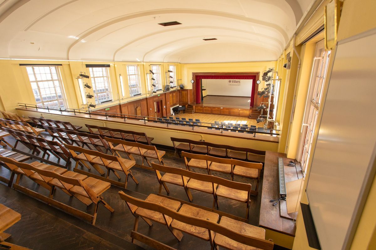 Empty auditorium with rows of wooden desks facing a stage and red curtains