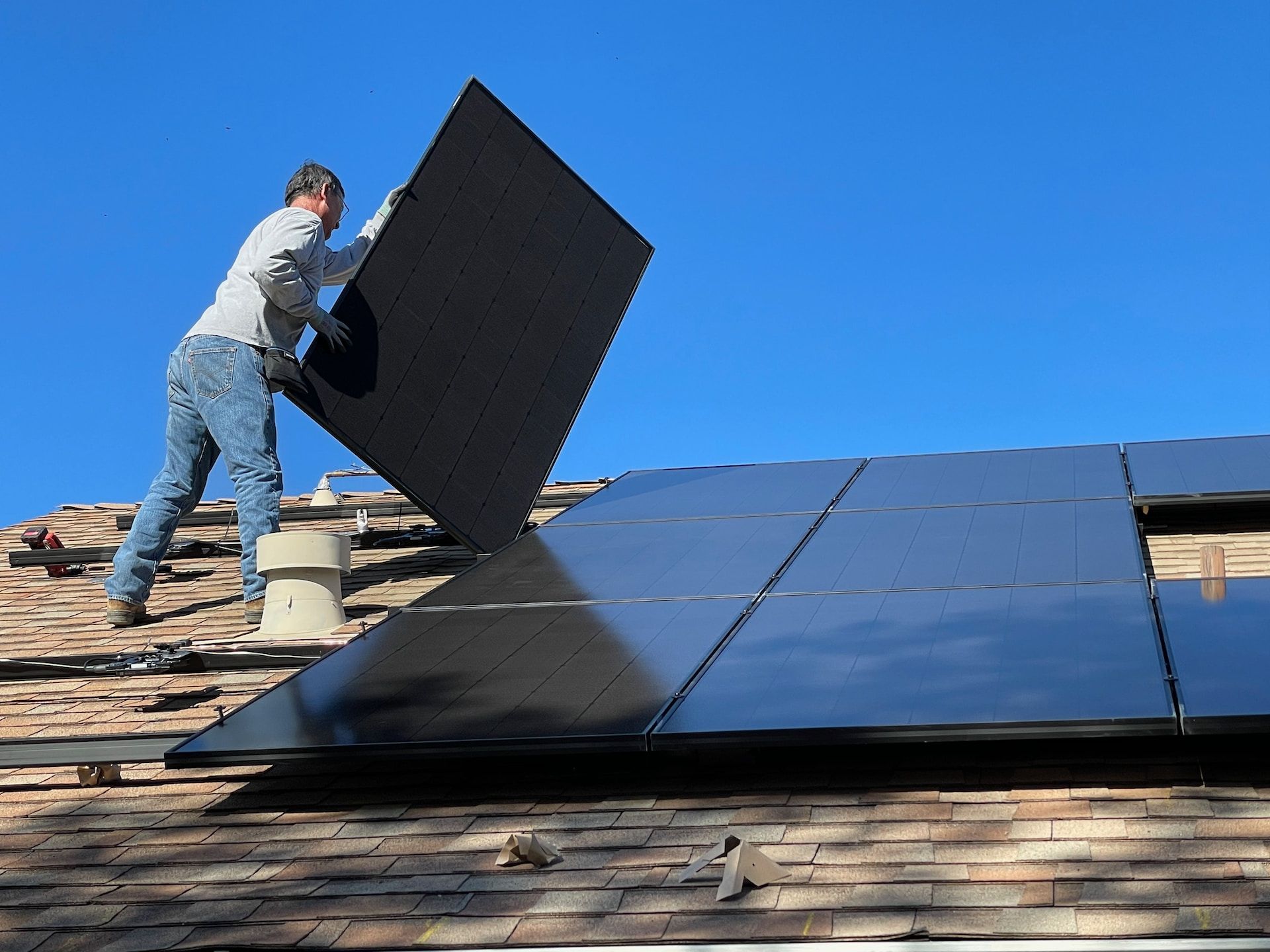 Worker installing black solar panels on a shingled roof under a clear blue sky