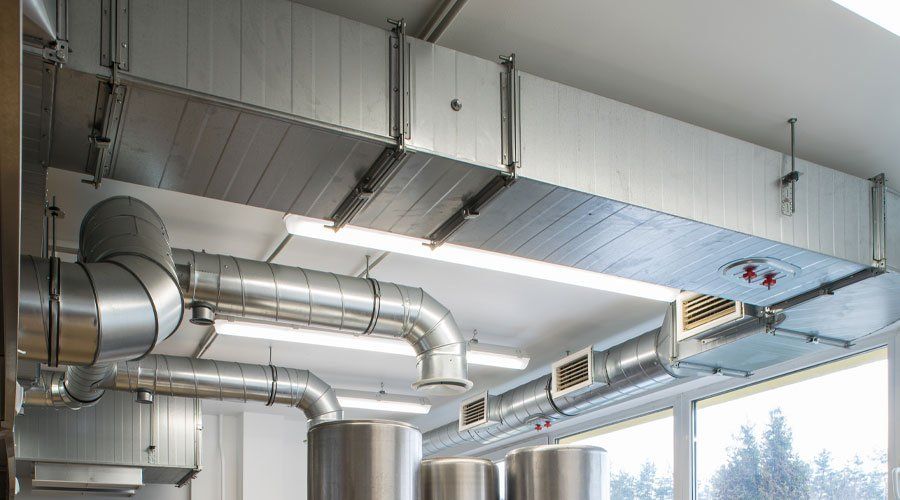 Industrial ceiling ductwork and vents in a bright interior room with large windows and exposed metal pipes.