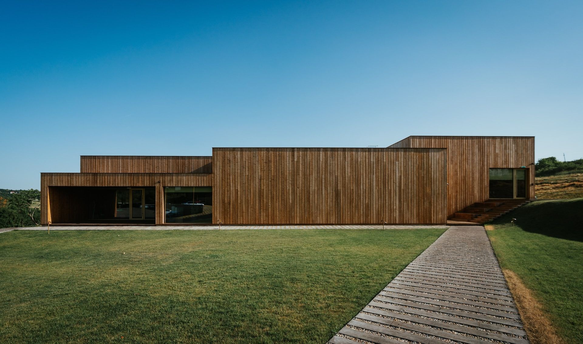 Modern wooden building on a grassy hill under a blue sky, with a paved walkway leading to the entrance