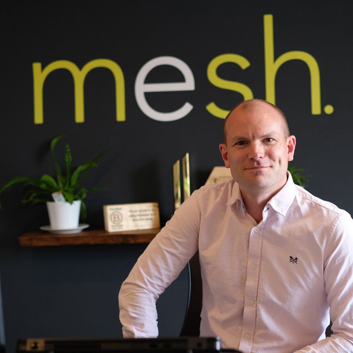 Man in a light pink shirt seated in an office with a black wall and large “mesh.” sign behind him