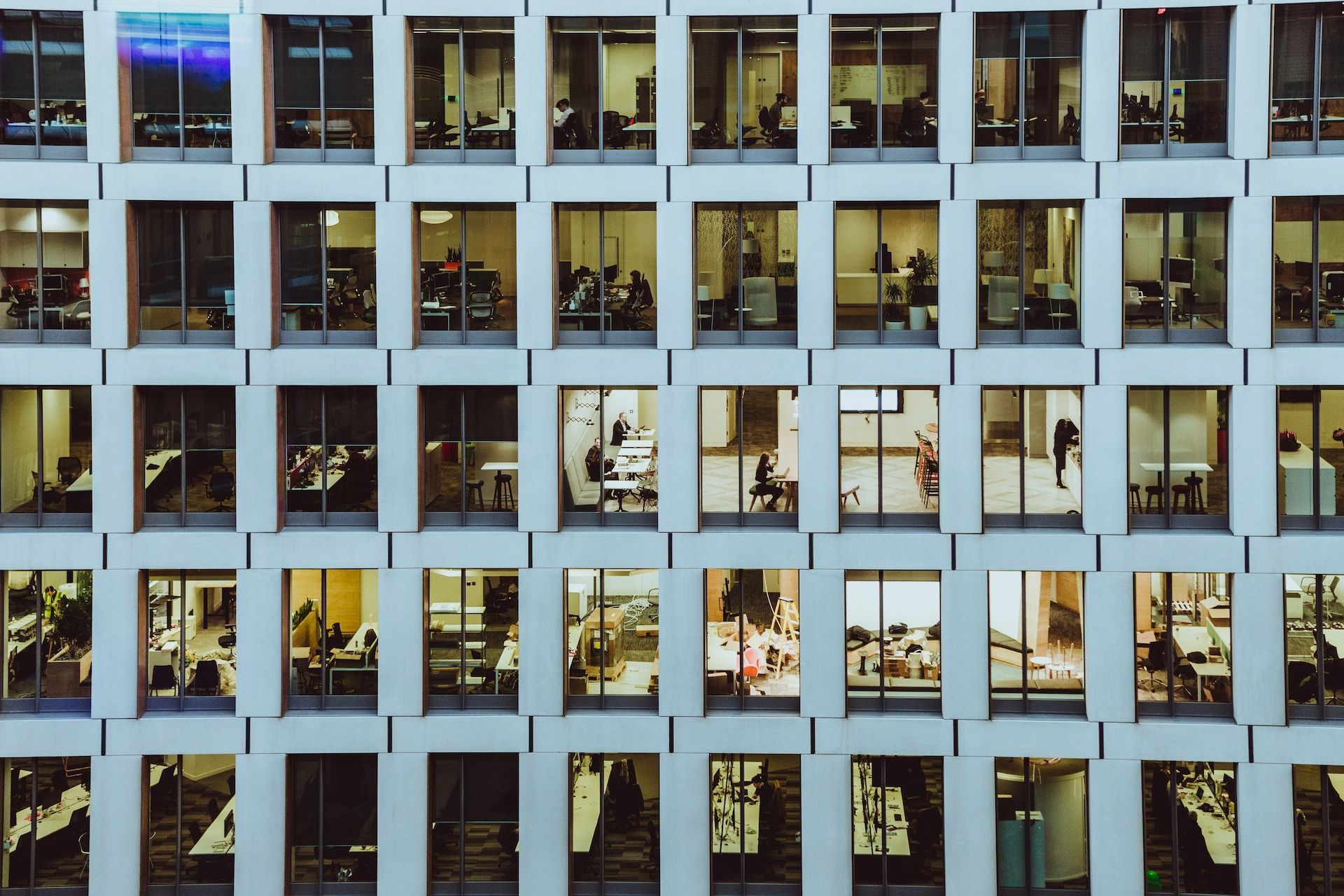 Office building façade with rows of lit windows and people working inside at dusk