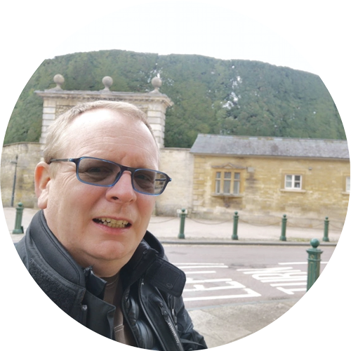 Man taking a selfie in front of a stone building and hillside, wearing sunglasses and a black jacket