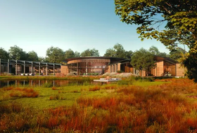 Modern building beside a grassy wetland under a clear sky, with trees and a wooden walkway