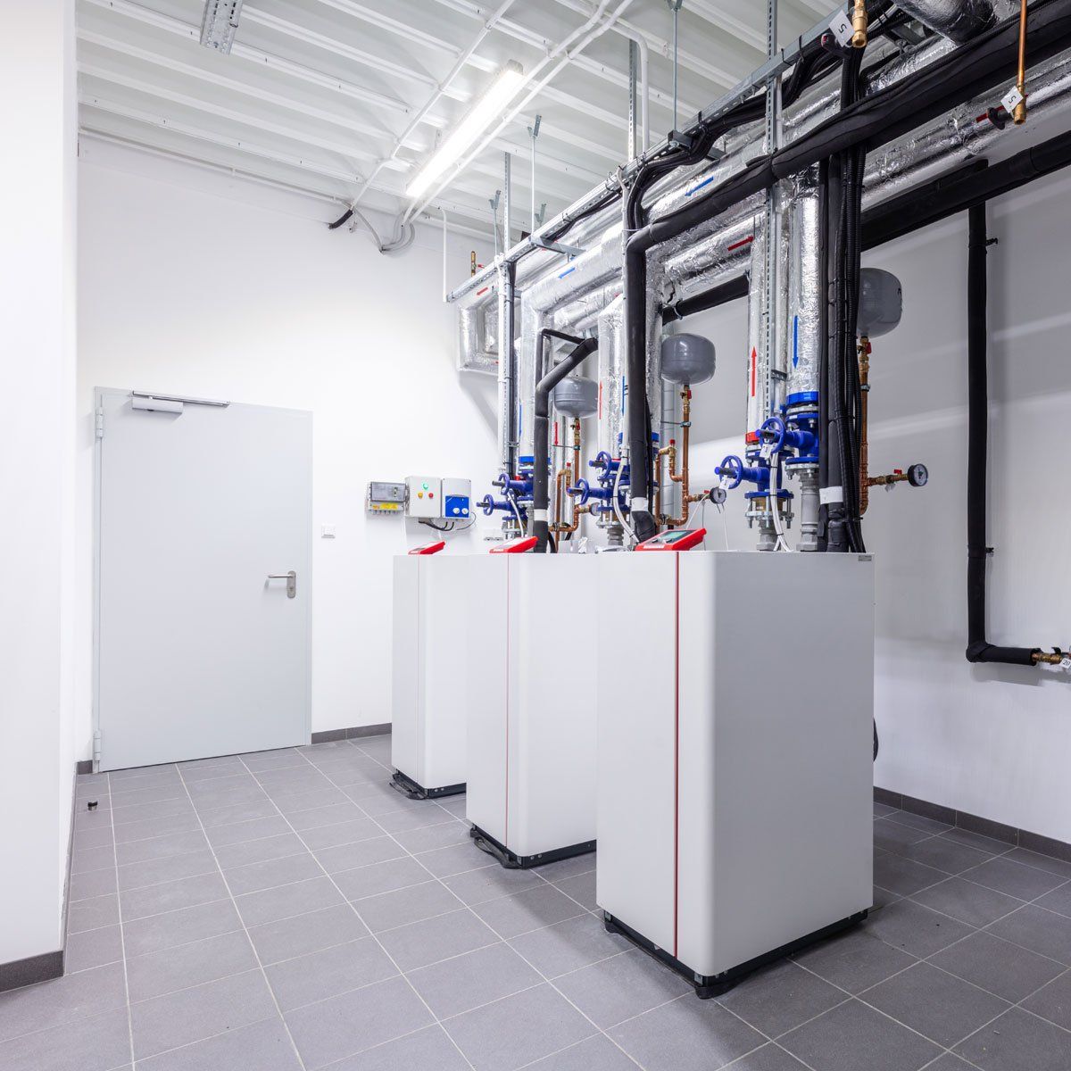 Bright utility room with white boilers, black pipes, and tiled floor under fluorescent lights