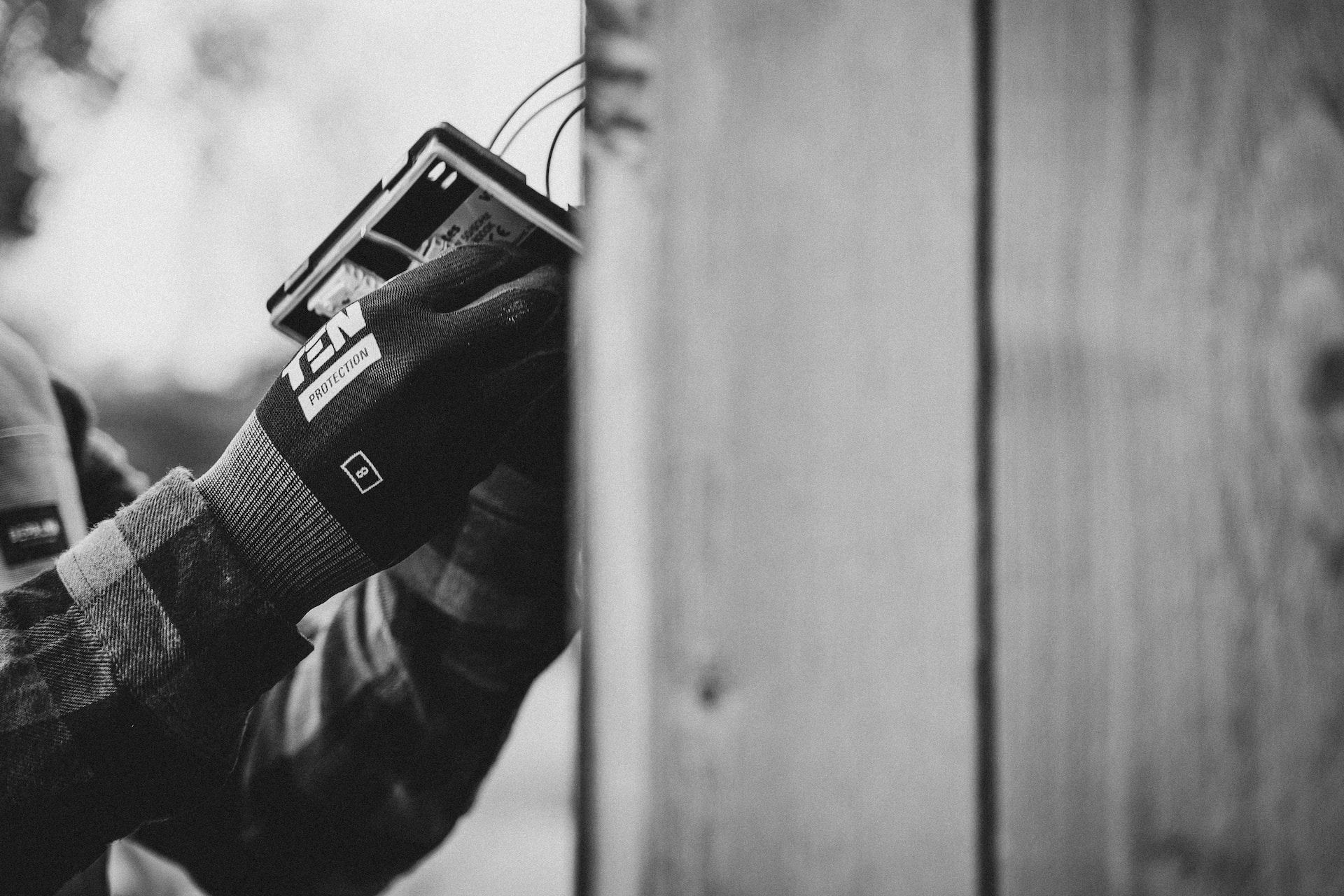 Gloved hands holding a small device beside a wooden wall in black-and-white close-up