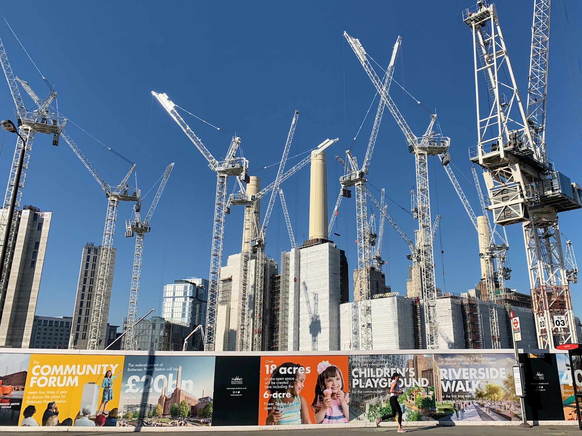 Construction cranes over high-rise buildings behind a colorful billboard fence under a clear blue sky