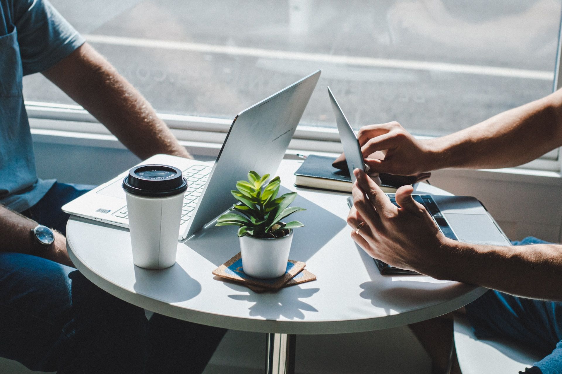 Two people working at a round table with a laptop, coffee cup, plant, and tablet by a bright window