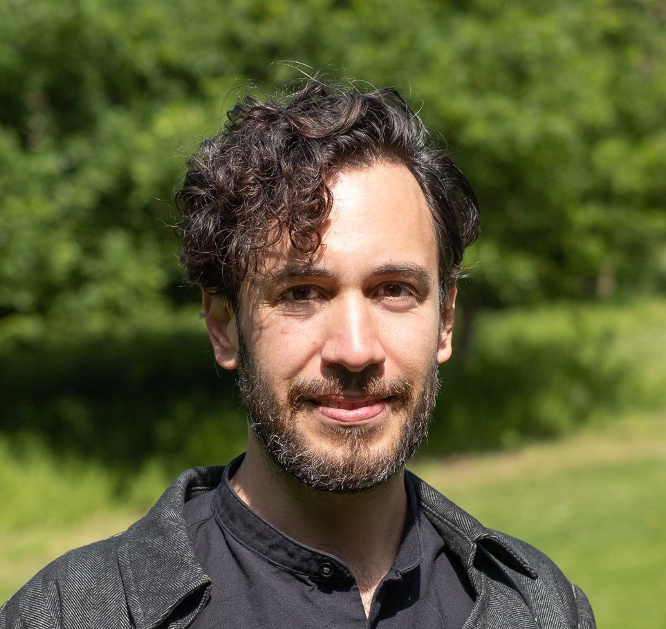 Portrait of a man with curly hair and a beard standing outdoors in a green park