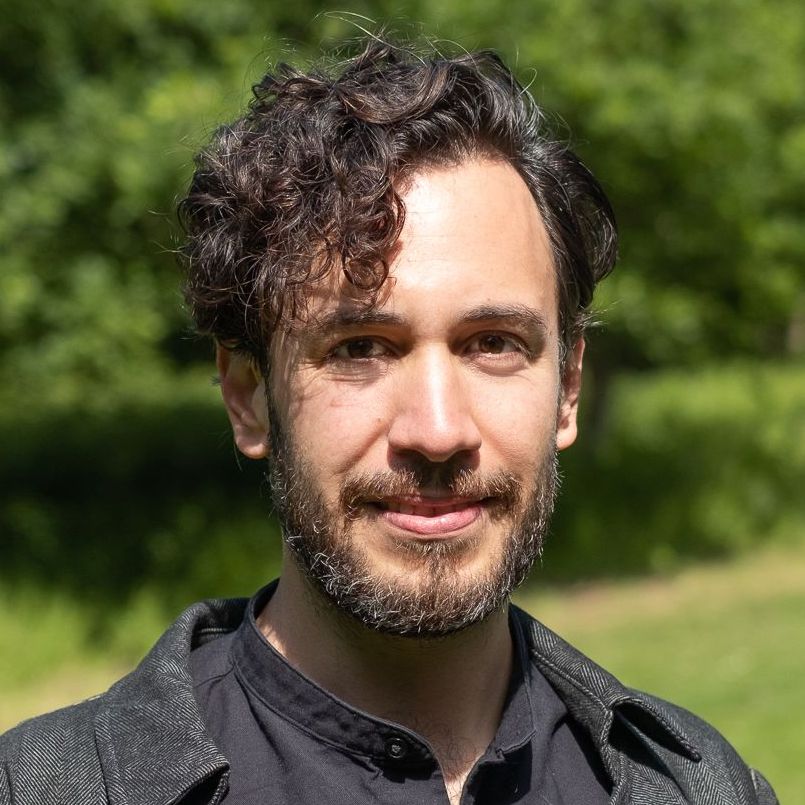 Smiling person outdoors in sunlight, with curly dark hair and a black shirt.